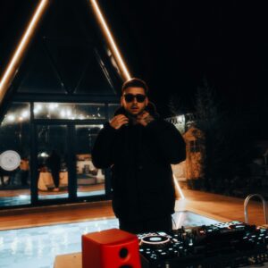 A DJ in sunglasses mixing tracks at a stylish nighttime pool party with vibrant lighting.